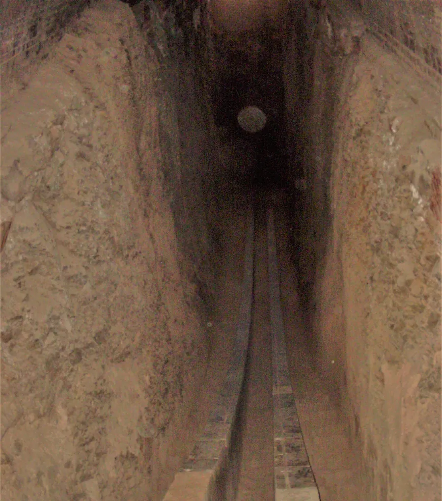 A deep, narrow stone-lined trench curving down into the ground, with two parallel rails along its floor